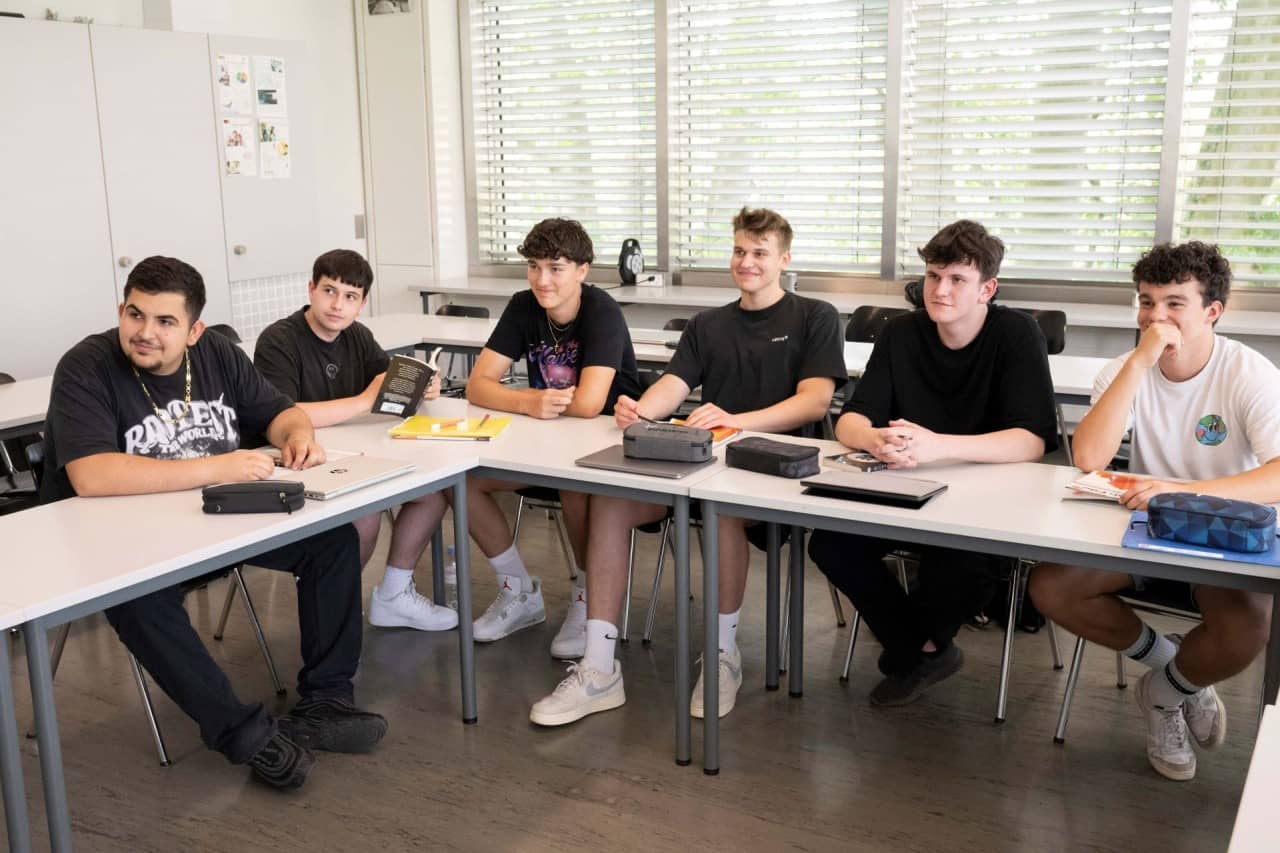 Sechs Schülerin in schwarzen T-Shirts sitzen an Schulbänken.