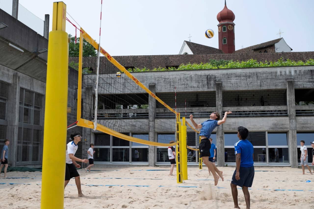Schüler auf dem Beach Volleyballfeld. Einer springt hoch um Volleyball übers Netz zu schlagen.