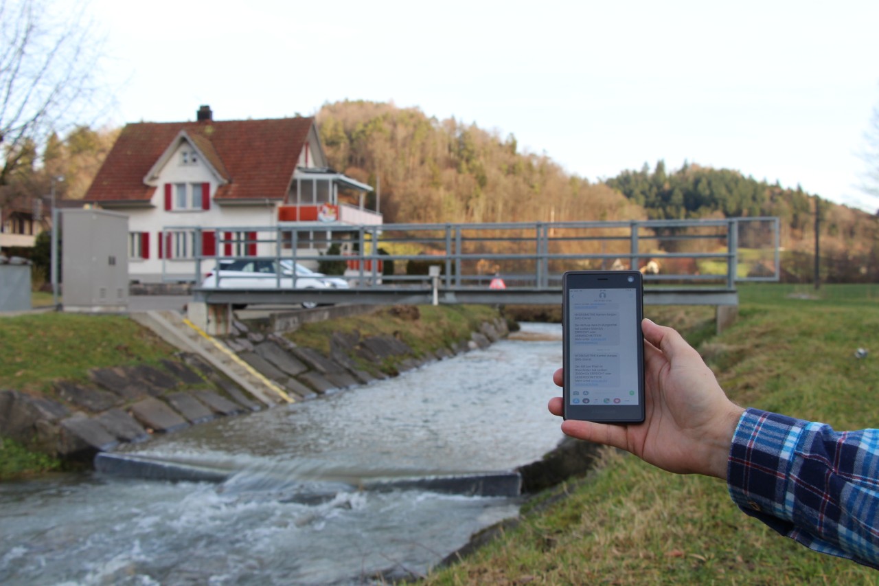 Eine Hand hält ein Smartphone auf dessen Bildschirm SMS zu sehen sind. Im Hintergrund ist eine Brücke über einen Bach zu sehen; an der Brücke ist eine Messstation installiert.