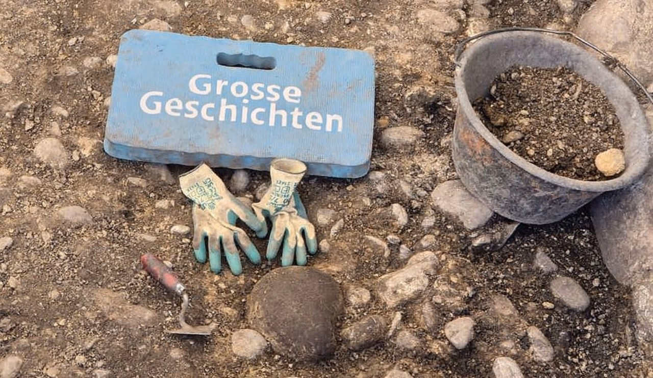 Ausgrabungsstilleben: links ein mit Erde gefüllter Kessel, daneben Handschuhe und eine Spitzkelle. Auf dem blauen Kniekissen steht "Grosse Geschichten".
