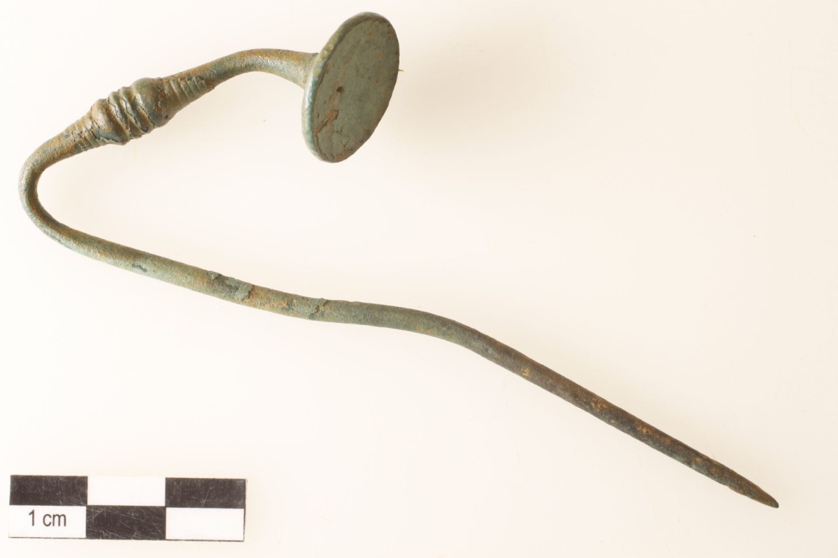 Hellgrün korrodierte Bronzenadel mit scheibenförmigem Kopf und Rippenverzierung im oberen Drittel. Verbogen.