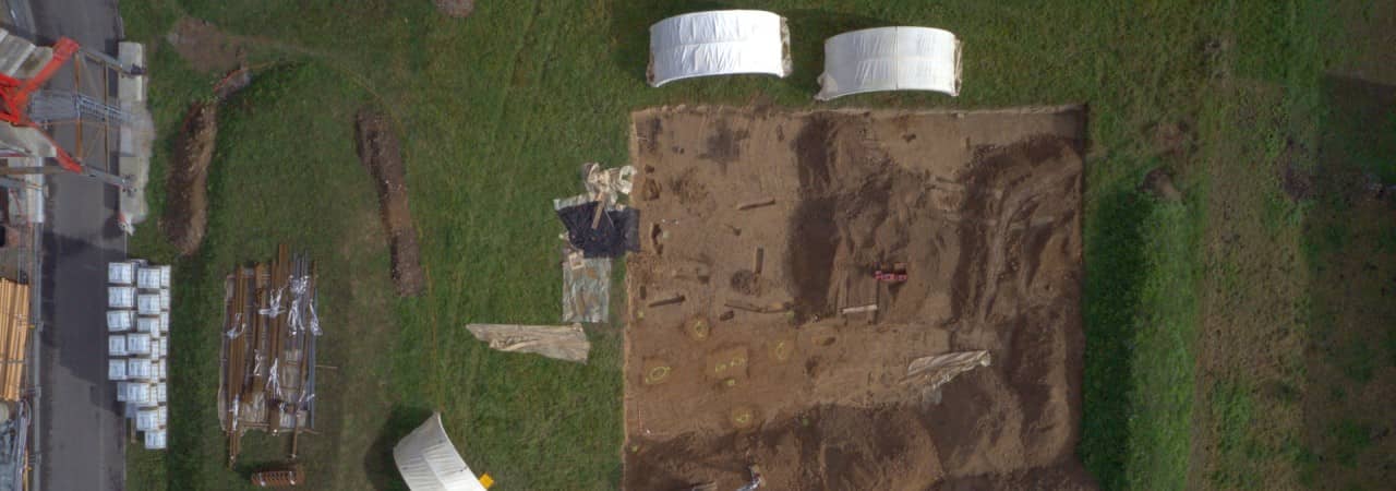 Drohnenbild der Ausgrabungsfläche inmitten deiner grünen Wiese. Am Rand oben stehen zwei weisse Grabungszelte, am unteren Bildrand sind Container und Autos aufgereiht.