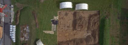 Drohnenbild der Ausgrabungsfläche inmitten deiner grünen Wiese. Am Rand oben stehen zwei weisse Grabungszelte, am unteren Bildrand sind Container und Autos aufgereiht.