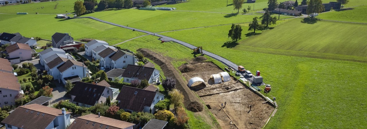 Drohnenaufnahme mit Ausgrabungsfläche im Vordergrund und klarer Weitsicht bis zu den Bergen.