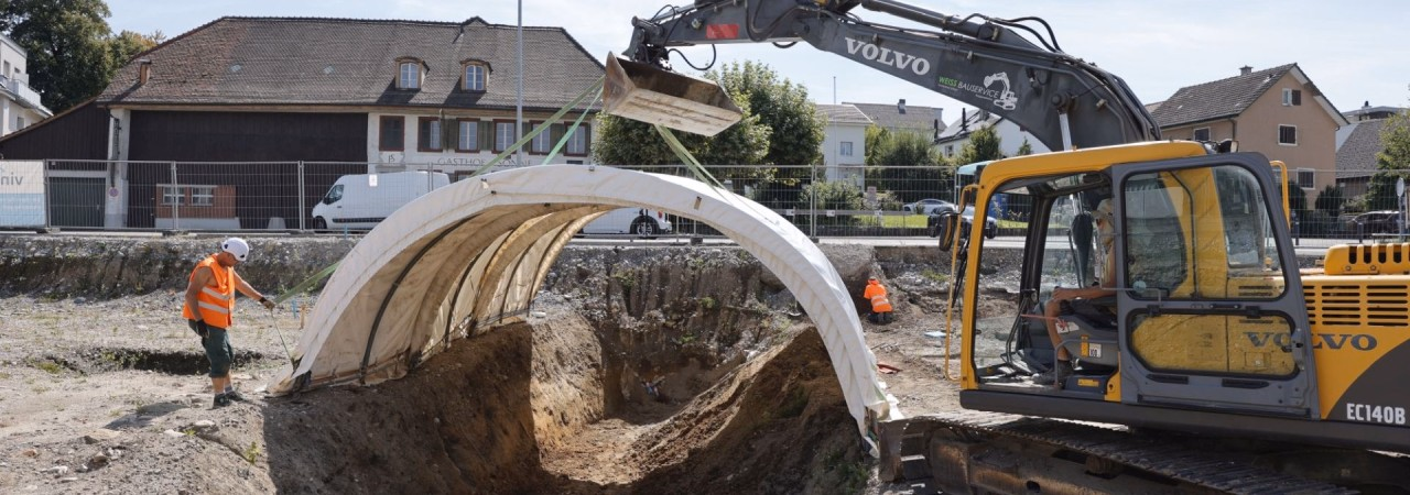Bagger stellt ein grosses Bogenzelt über einen Ausgrabungsschnitt hin.