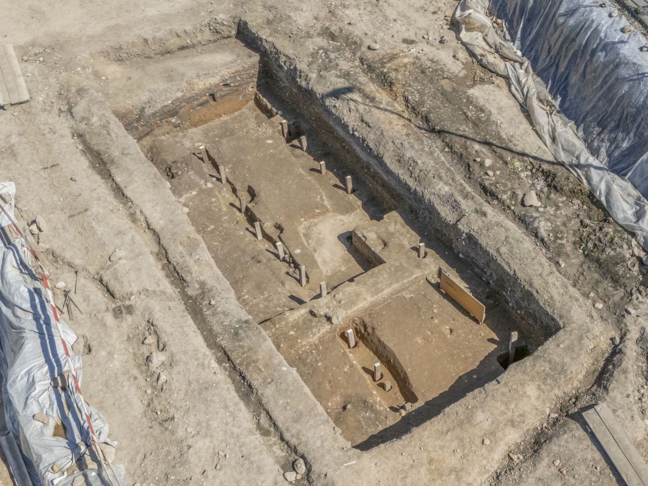 Archäologische Ausgrabungsstätte mit rechteckigen Strukturen und freigelegtem Erdreich, umgeben von Abdeckplanen und Werkzeugen.