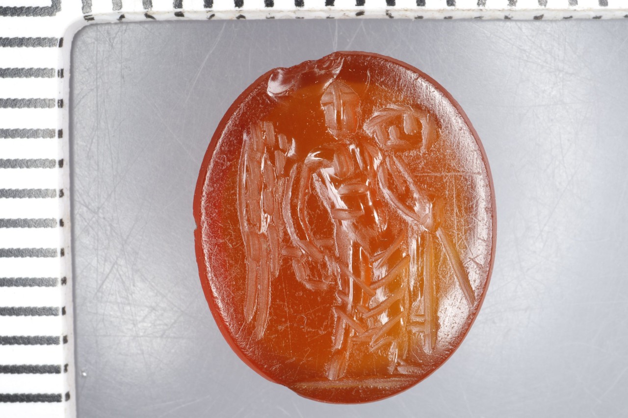 Roter Schmuckstein mit eingravierter Figur, eine stehende Person in Rüstung mit Stab und Kranz.