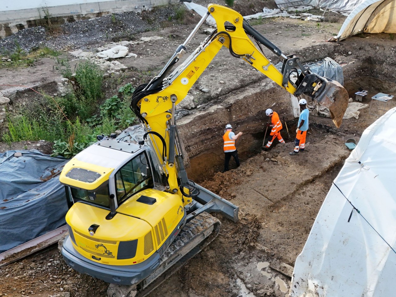 Ein gelber Bagger hebt Erde aus einer archäologischen Grabungsstätte, daneben drei Personen in Arbeitskleidung mit Helmen, die am Profil arbeiten.