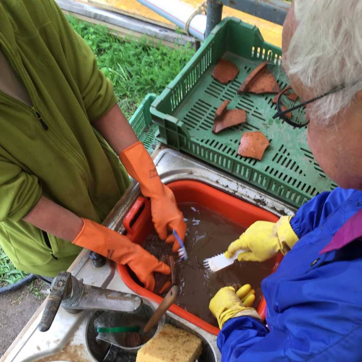 Blick von oben auf ein Waschbecken, an dem zwei Personen mit farbigen Gummihandschuhen Scherben waschen.