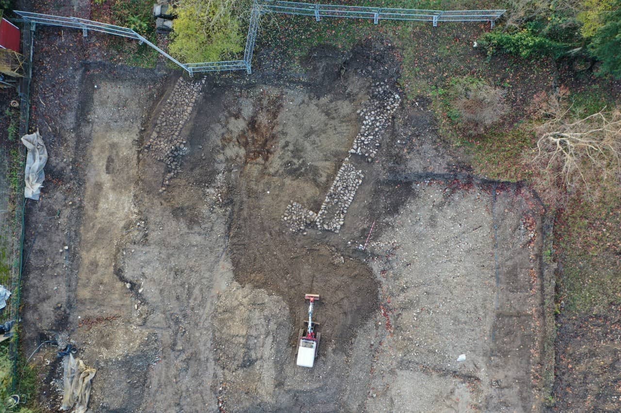 Drohnenaufnahme der Grabungsfläche mit Bagger am unteren Bildrand und freigelegten Fundamenten der Kirche am oberen Bildrand.