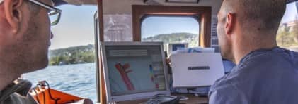 Zwei Experten betrachten die Messaufnahmen der Welterbestätte an einem Bildschirm auf einem Boot.