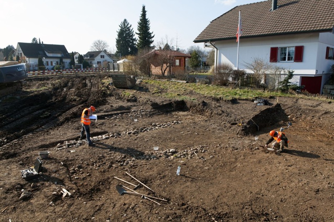 Ein Ausgräber legt rechts im Bild Strukturen frei und ein Ausgräber zeichnet links im Bild die Befunde. 