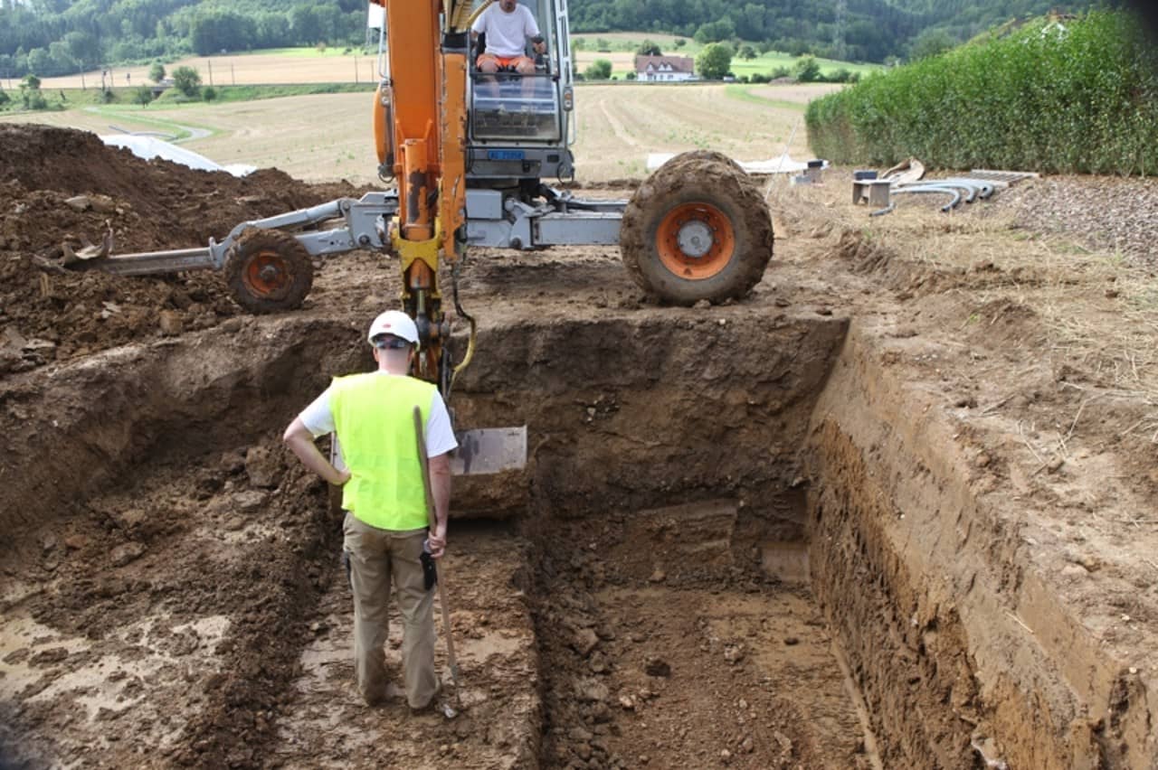 Archäologe im Vordergrund und Bagger in Hintergrund.