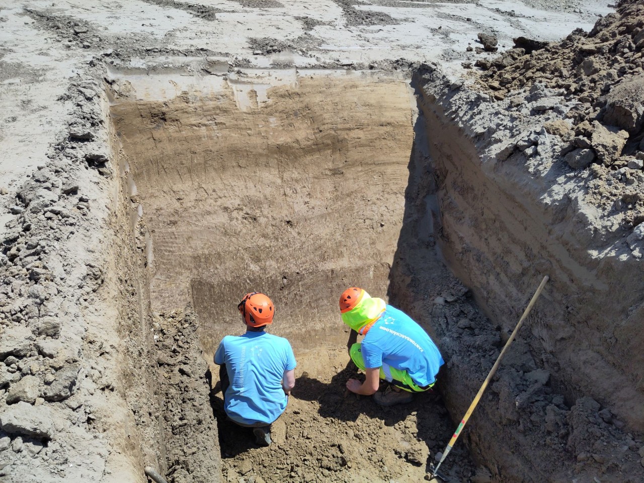 Zwei Grabungsmitarbeiter mit Sicherheitshelmen bei der Untersuchung einer tiefen Erdgrube auf einer Baustelle.