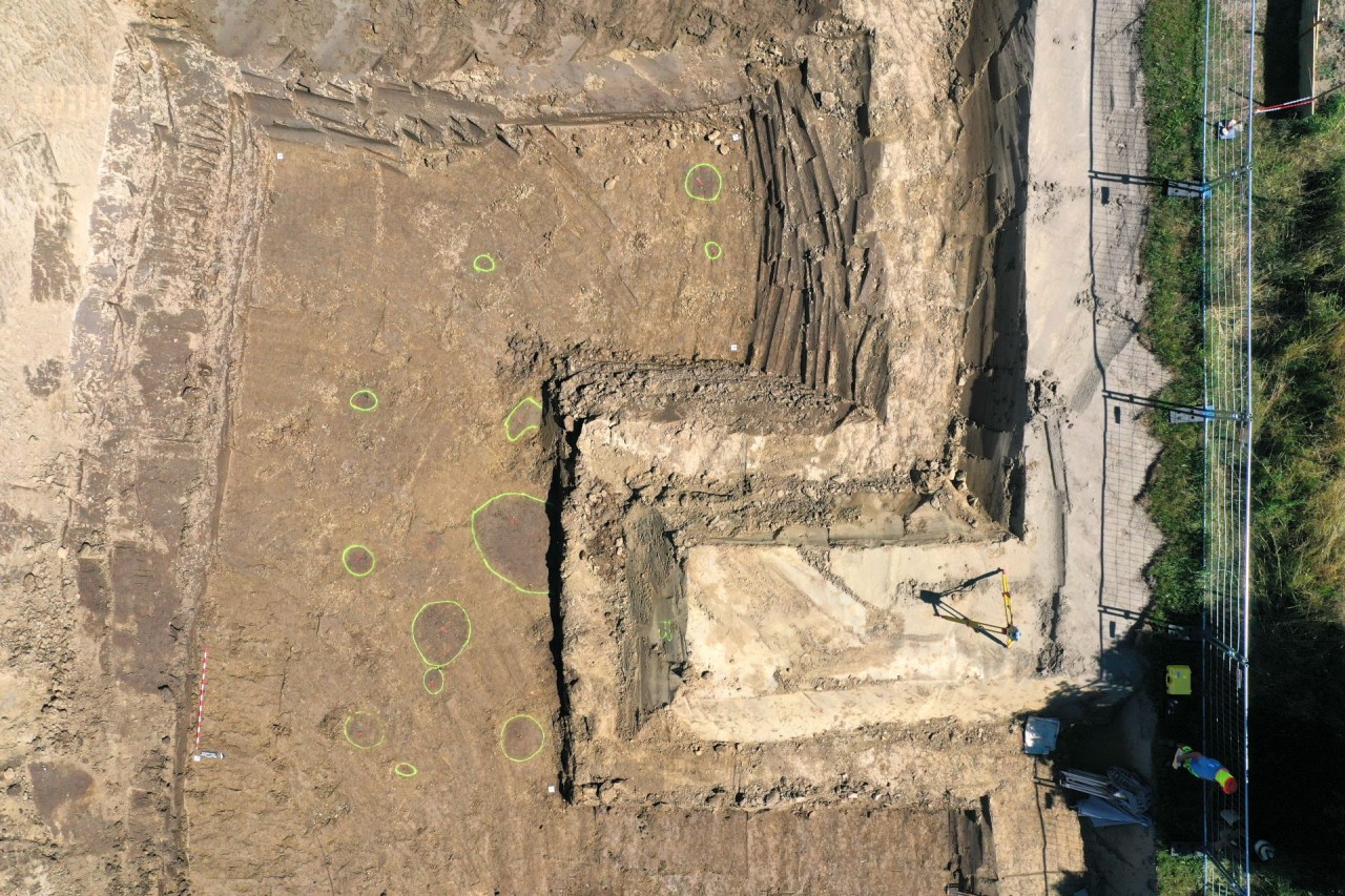 Luftaufnahme einer archäologischen Grabungsstätte mit eingezeichneten Kreisen im Boden, rechteckigen Ausgrabungsgrenzen und zwei Arbeitenden am rechten Bildrand.