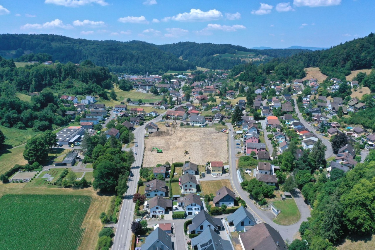 Luftaufnahme einer Ortschaft in hügeliger Landschaft mit Häusern, Straßen und einem freien Baugelände in der Mitte. Im Hintergrund bewaldete Hügel unter blauem Himmel.