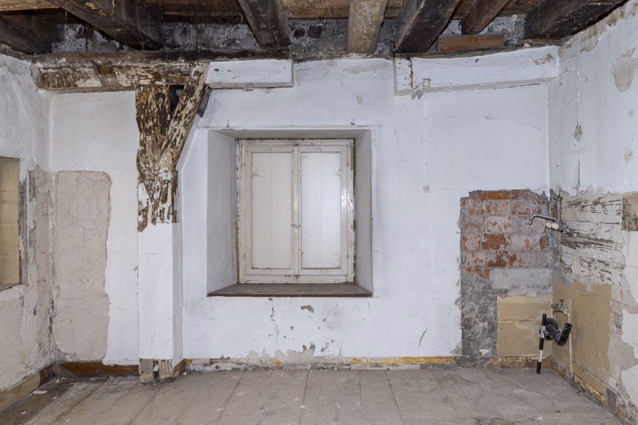 Anblick einer weissgestrichenen Binnenwand mit links einem Pfosten auf übermalten Holz und im Zentrum ein Fenster.