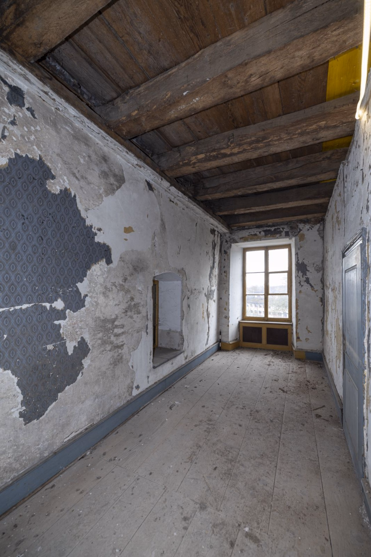 Blick in ein enges Zimmer, rechts der Rest von blauer Tapete, oben Holzdecke und hinten Blick aus dem Fenster.