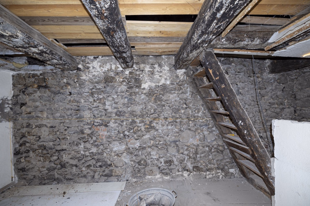 Blick auf eine gemauerte Wand aus Bollensteinen, oben die hölzerne Decke und rechts eine Treppe, die ins obere Geschoss führt.