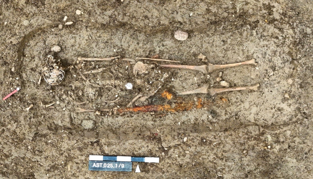 Freigelegtes Skelett in einer Grabgrube. An der Seite liegt ein braun korrodiertes Eisenschwert.