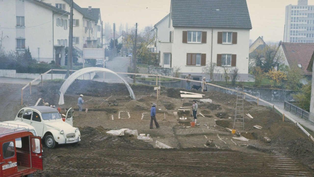 Auf einer Baustelle stehen fünf Personen zwischen Erdhügeln und Bauwerkzeugen. Im Vordergrund ein weißes Auto und ein rotes Fahrzeug. Im Hintergrund Wohnhäuser sowie ein großes Gebäude.