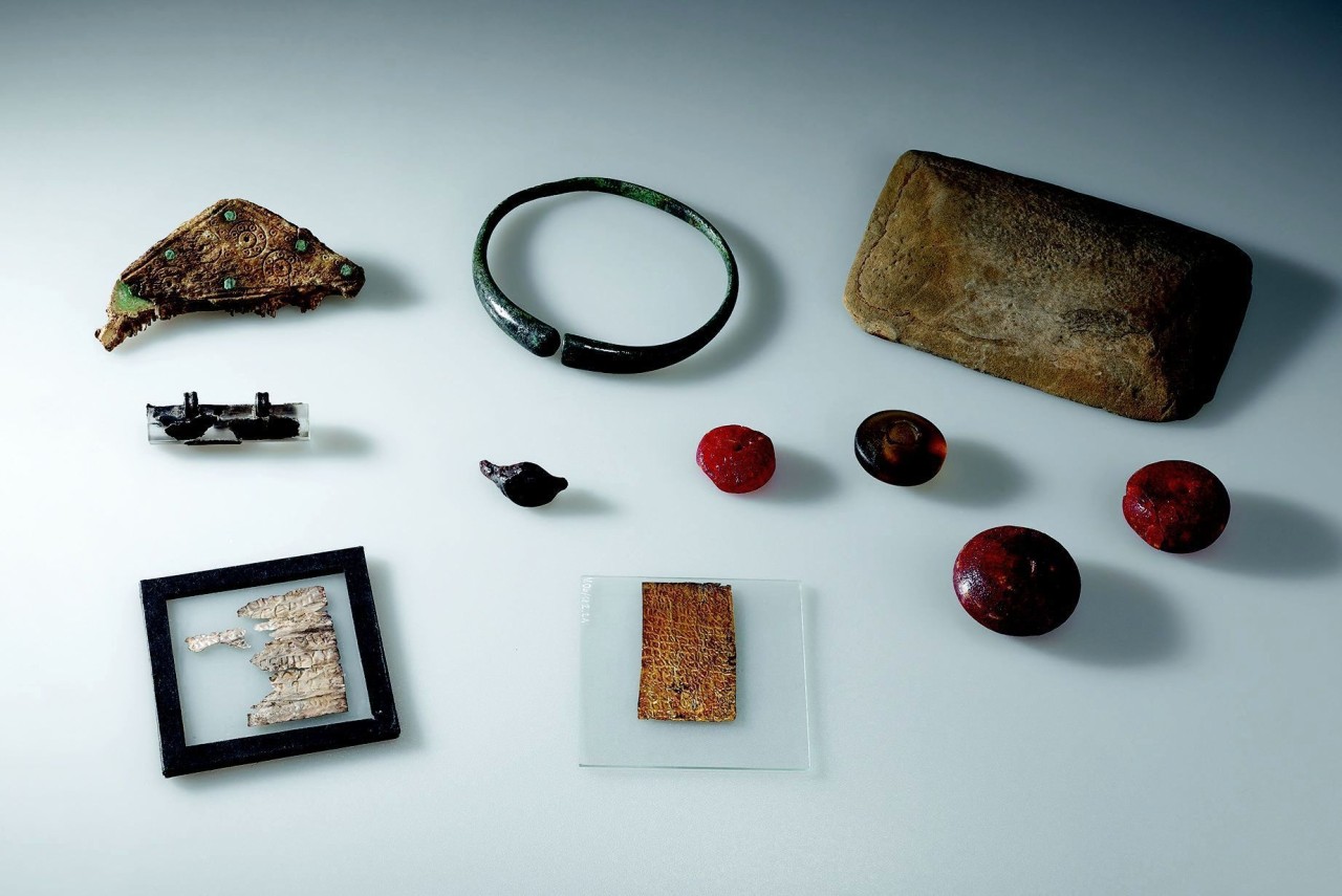 Archäologische Fundstücke, darunter ein Bronzeschmuck, ein Armreif, eine Steintafel, rote Schmucksteine sowie ein Gold- und Silberblech mit Schriftzeichen zwischen Glasplatten, auf einer hellen Oberfläche arrangiert.