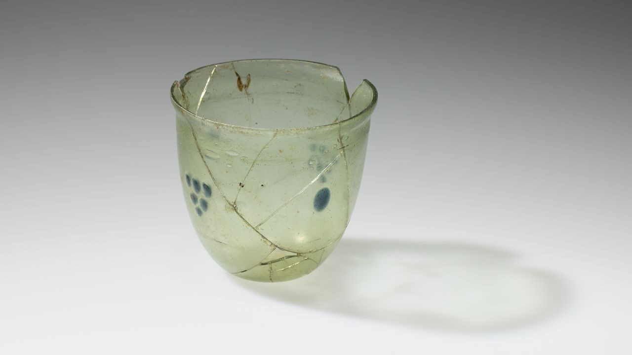 Ein beschädigtes, hellgrünes Glasgefäß mit mehreren Bruchstellen und sichtbaren Reparaturstellen. Links sind blaue Musterpunkte, rechts ein einzelner blauer Punkt. Das Gefäß wirft einen Schatten.