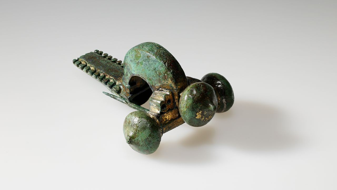 Fibel aus grünlicher Bronze mit bogenförmigem Kopf und kugelförmigen Verzierungen, auf hellem Hintergrund.