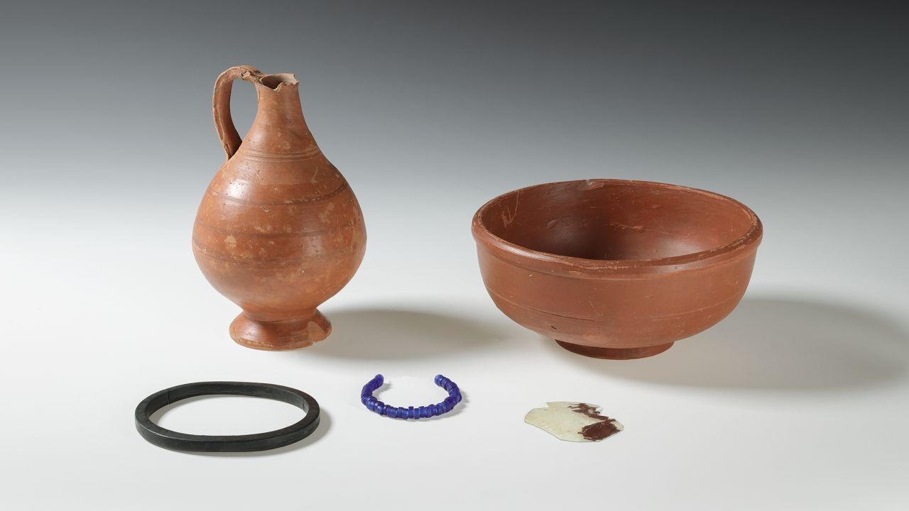 Keramikkrug und Schale aus rotem Ton, daneben drei kleine Objekte: ein schwarzer Ring, eine blaue Perlenkette und ein Glasfragment mit braunen Stellen.