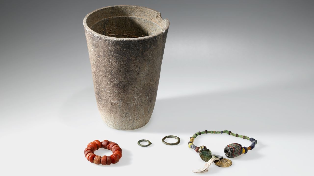 Ein Becher aus Lavez neben archäologischen Funden: ein rotes Perlenarmband, zwei Metallringe, eine mehrfarbige Halskette und Anhänger aus verschiedenen Materialien.