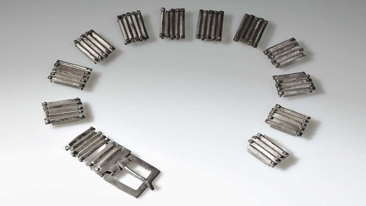 Metallglieder eines silbernen Gürtelverschlusses in halbkreisförmiger Anordnung auf hellem Hintergrund.