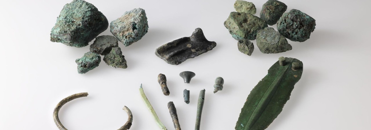 Mehrere Bronzeobjekte als Ensemble angeordnet: Fragmente von Barren, ein Bronzedolch, Fragmente von Nadeln, ein Sichelfragment und ein Armring.