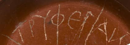Fragment eines roten Keramiktellers mit eingeritzter Inschrift in griechiscgen Buchstaben.