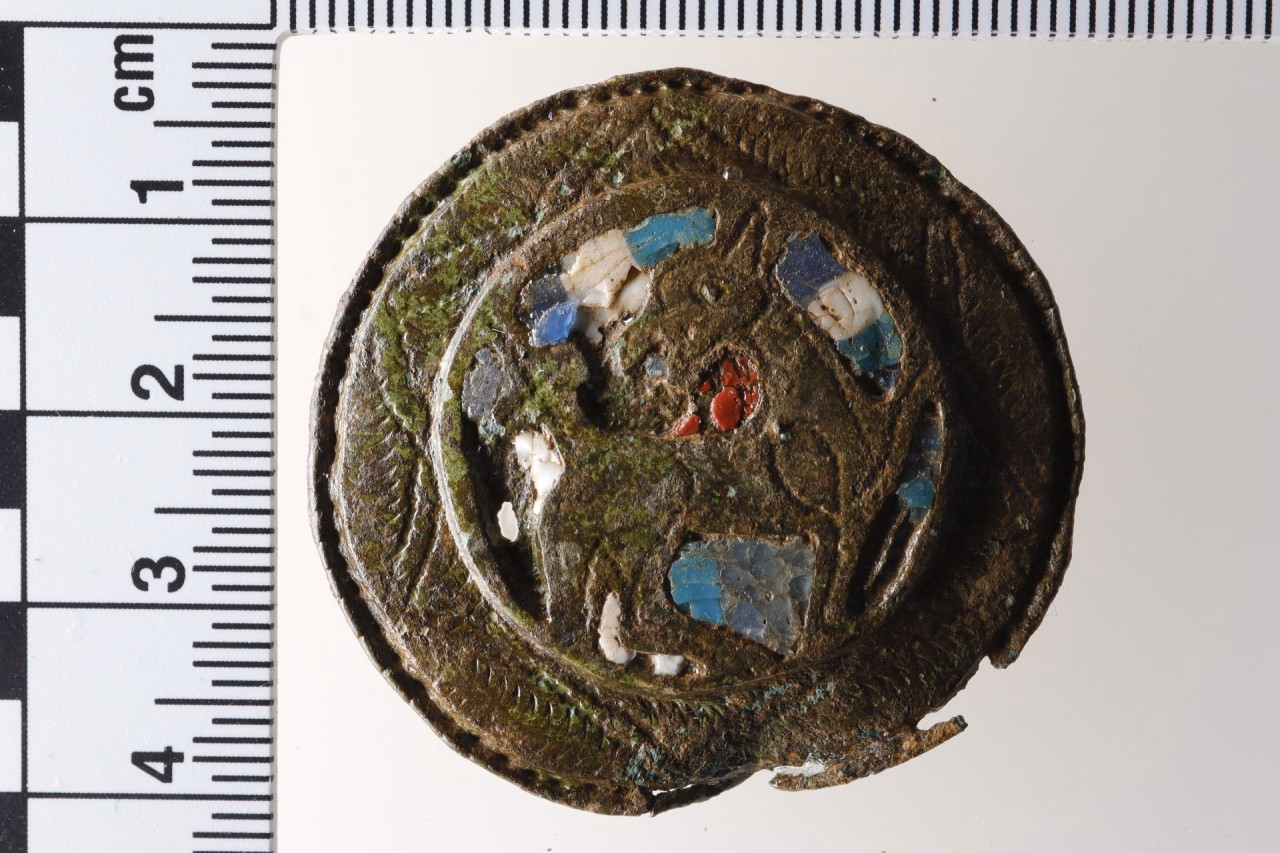 Runde Fibel aus Bronze, mit farbigen Einlagen aus Glas. Eingraviert ist ein stilisiuertes Lamm.