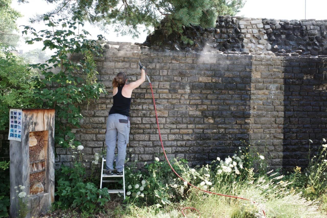 Eine Frau reinigt mit einem Gerät die römische Mauer. Rechts ist ein heller Streifen zu sehen
