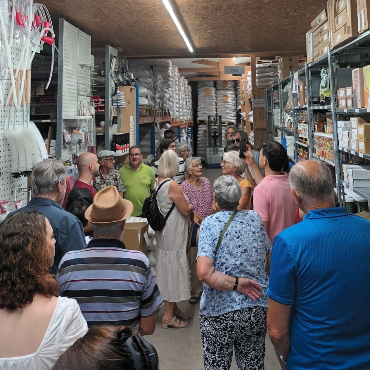 Eine Gruppe von etwa 15 Personen steht in einem Lagerraum von Brau und Rauch mit Regalen, Kartons und Waren. Die Teilnehmer schauen und hören einer Person zu, die etwas erklärt.