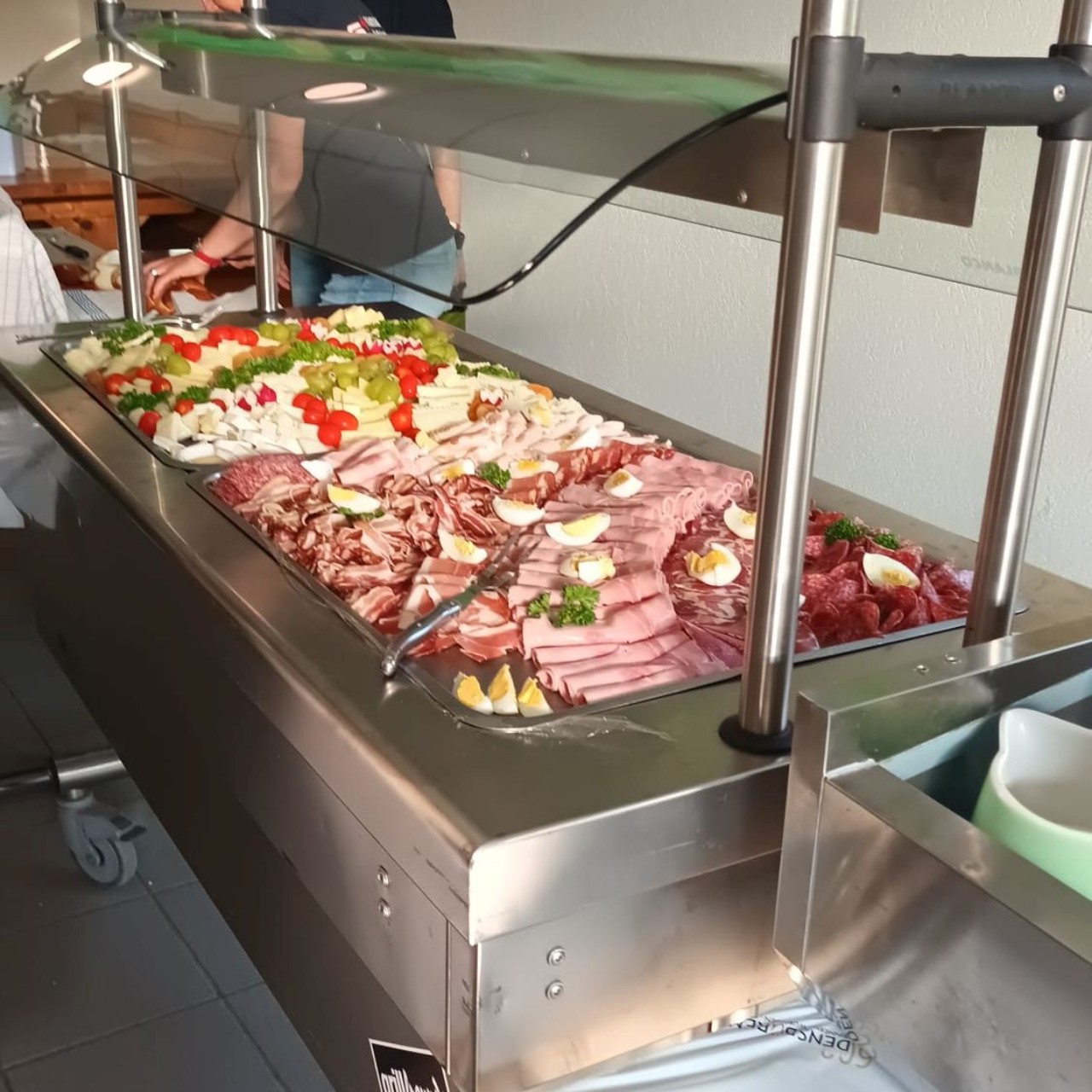 Buffet mit kalten Speisen, darunter Wurst, Schinken, Käse, Tomaten, Salat und hartgekochte Eier, angerichtet auf einer Edelstahl-Salatbar.
