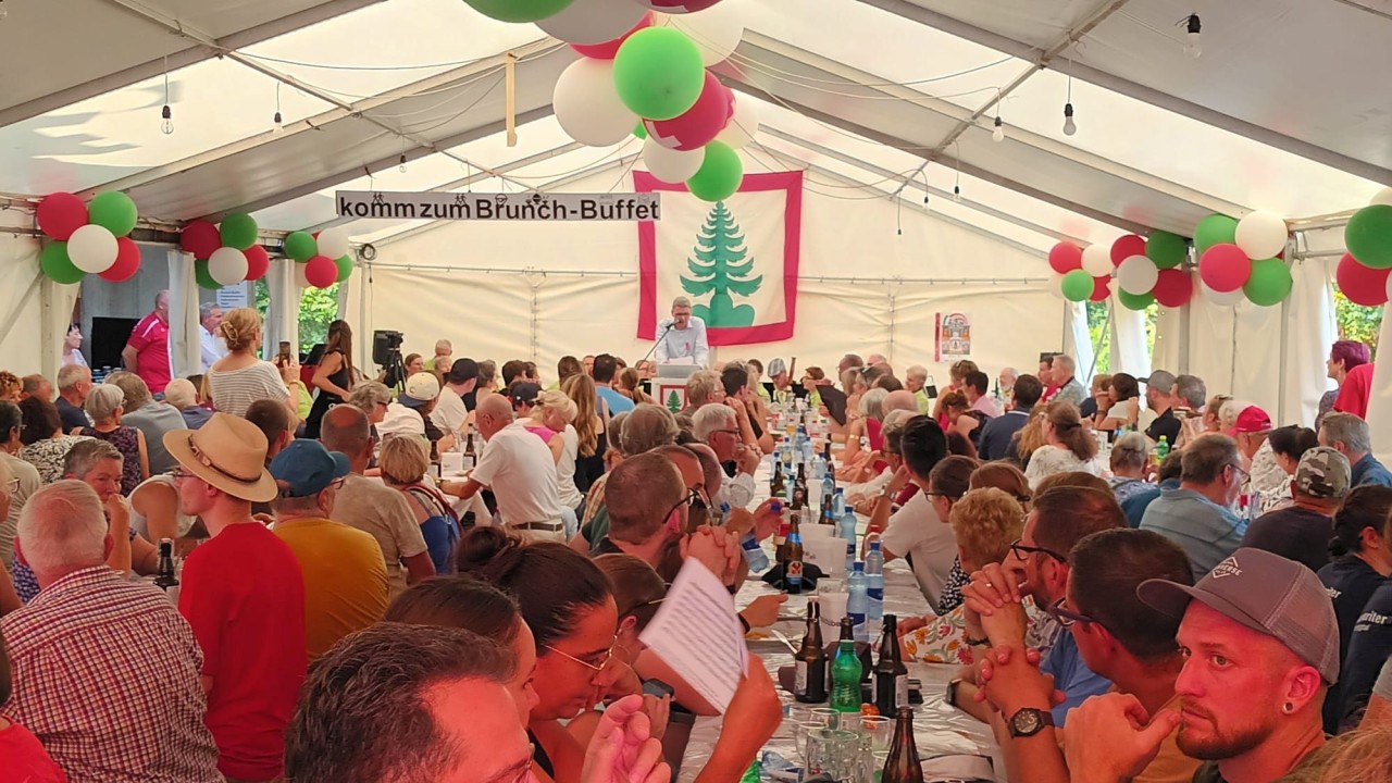 Ein gut besuchtes Festzelt mit Menschen an langen Tischen, geschmückt mit Ballons in Rot und Grün. Im Hintergrund Regierungsrat Dieter Egli am Rednerpult sowie ein Banner mit dem Wappen von Densbüren, einem grünen Tanne. Text: "komm zum Brunch-Buffet".