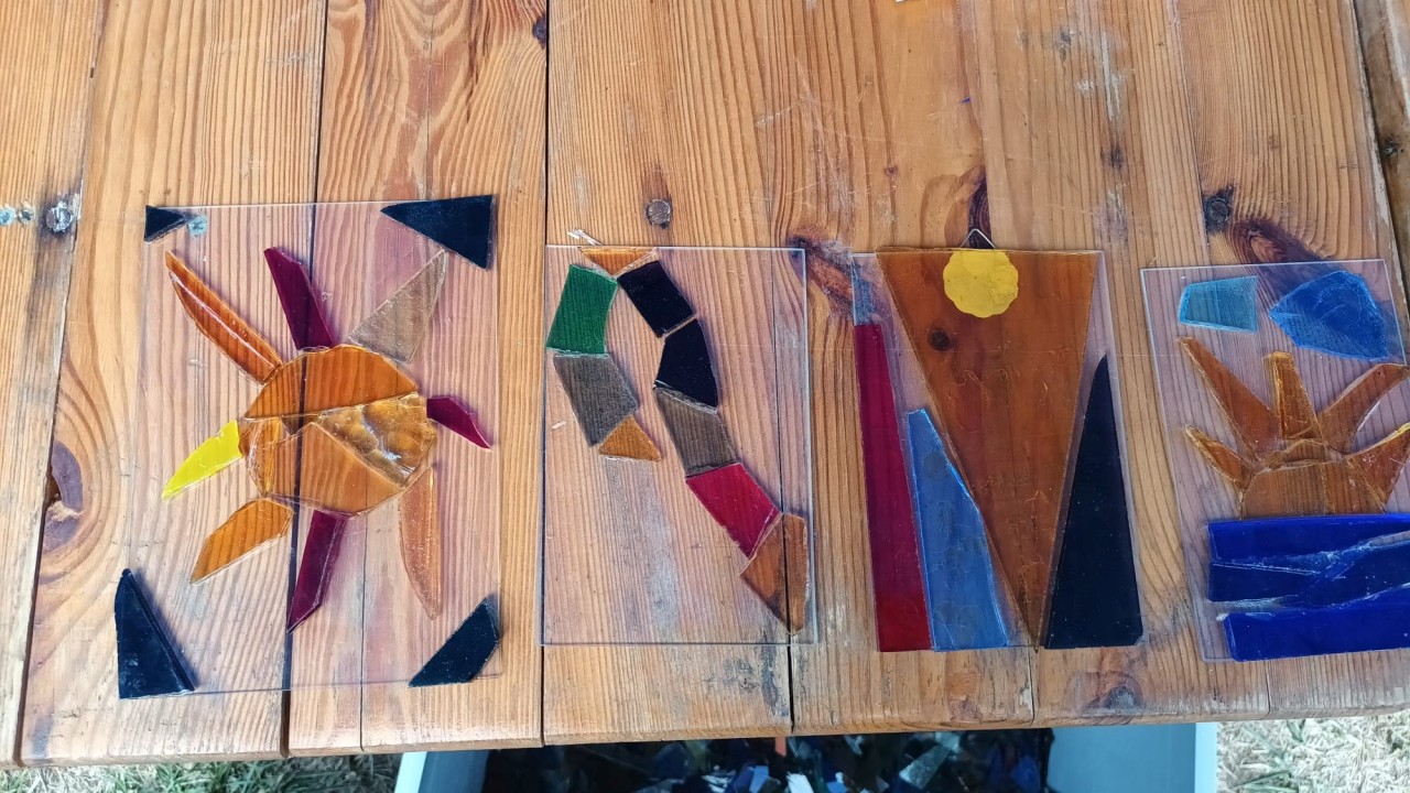 Farbig gestaltete Mosaike aus Glas auf einem Holztisch, bestehend aus Formen wie abstrakten Mustern und einer Sonne über Wasser.