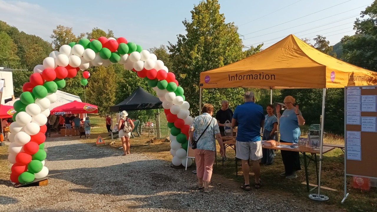 Ein bunter Luftballonbogen in Grün, Weiß und Rot vor einem gelben Infozelt mit Personen. Links weitere Zelte und Bäume im Hintergrund.