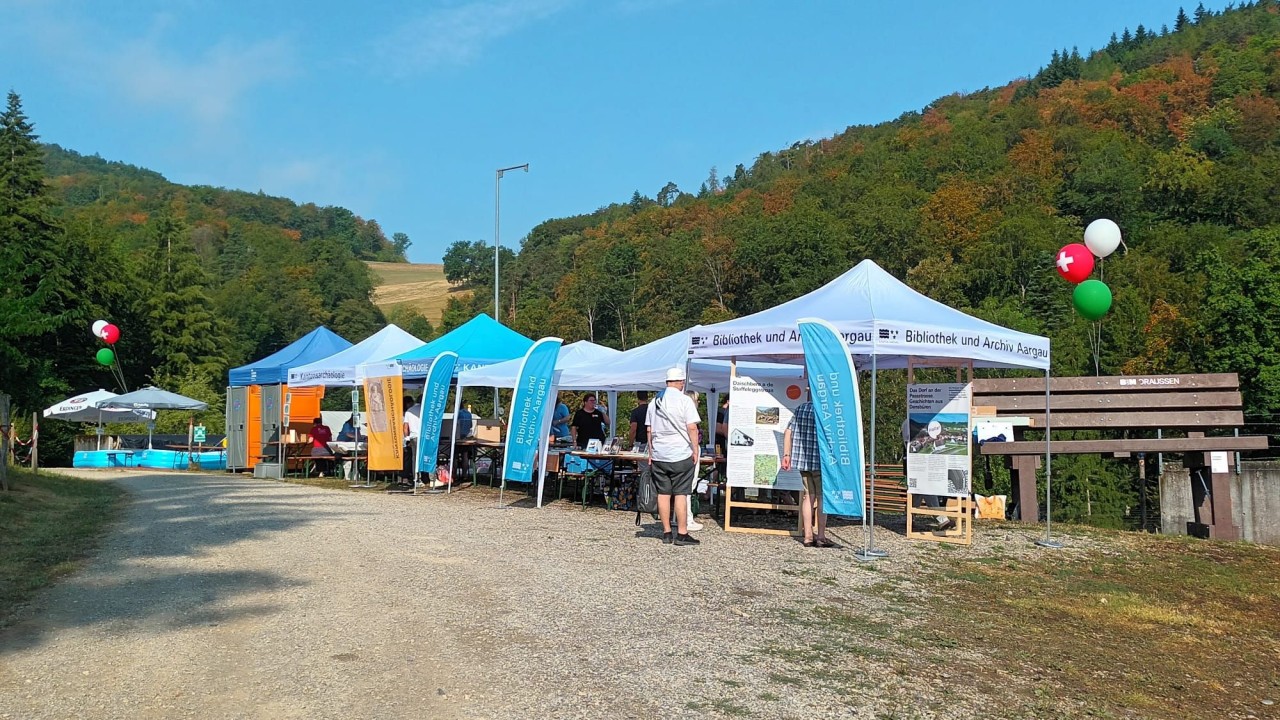 Mehrere Pavillons mit Infoständen und Rollups auf einem Kiesweg vor einer bewaldeten Hügelkulisse, Luftballons an den Zelten befestigt, Personen stehen und lesen Informationsmaterial.