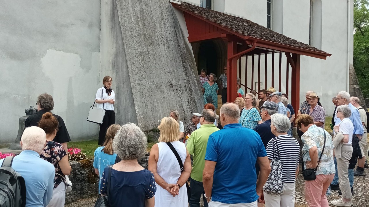 Eine Gruppe von Menschen bei einer Führung vor der Kirche mit weiss-grauer Fassade und rotem Holzdach.
