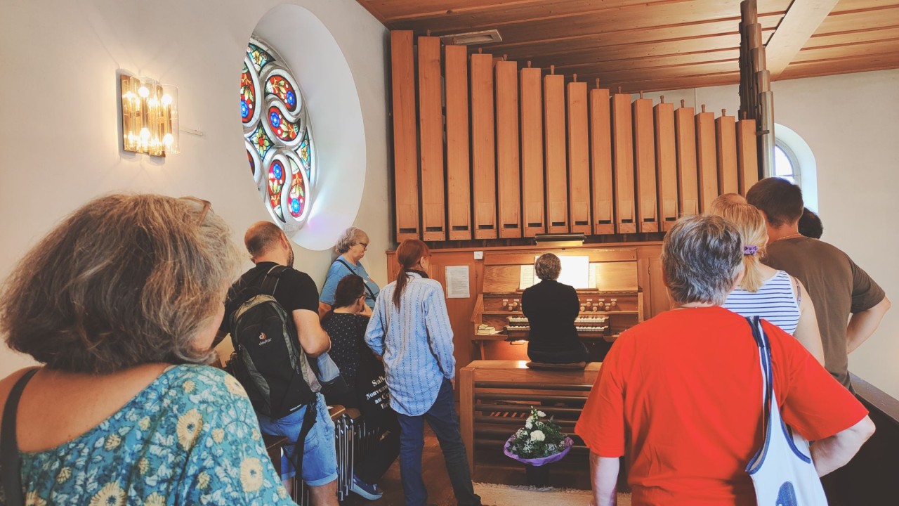 Mehrere Personen, die eine Orgel in einem Raum mit Holzdecke und farbigem Rundfenster betrachten, eine Person spielt die Orgel.