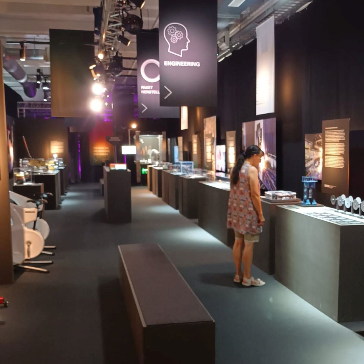 Eine Person betrachtet Exponate in einem modernen Ausstellungsraum mit Tischen, erklärenden Tafeln und Beleuchtung. Oben hängen Schilder mit Informationen und Symbolen.