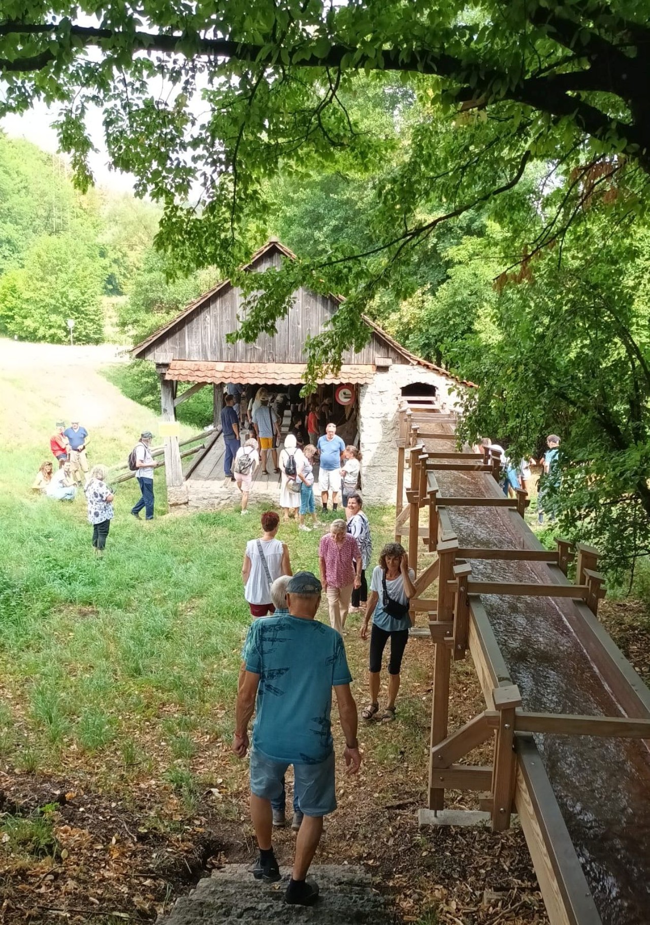 Eine Gruppe von Menschen vor einer historischen Säge mit hölzerner Wasserrinne inmitten einer grünen Landschaft.