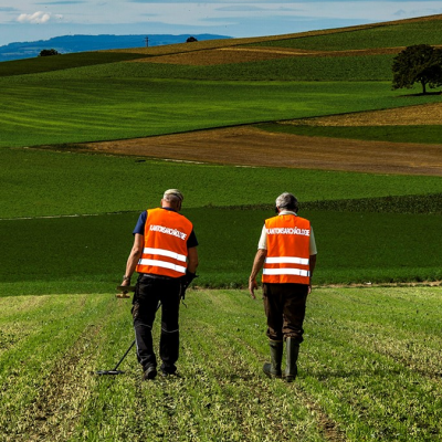 Vier Metalldetektorgänger in orangen Leuchtwesten untersuchen ein frisch gemähtes Feld.