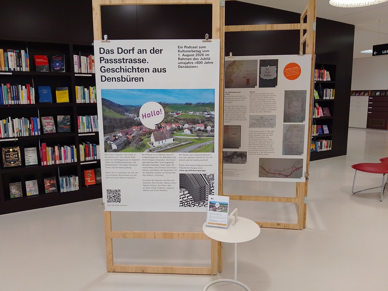 Ausstellung mit zwei Stellwänden, Büchern im Hintergrund. Links Plakat mit Titel 'Das Dorf an der Passstrasse' über das Dorf Densbüren. Rechts historische Informationen, darunter Karten und Bilder. Vordergrund: runder Tisch mit Broschüren.