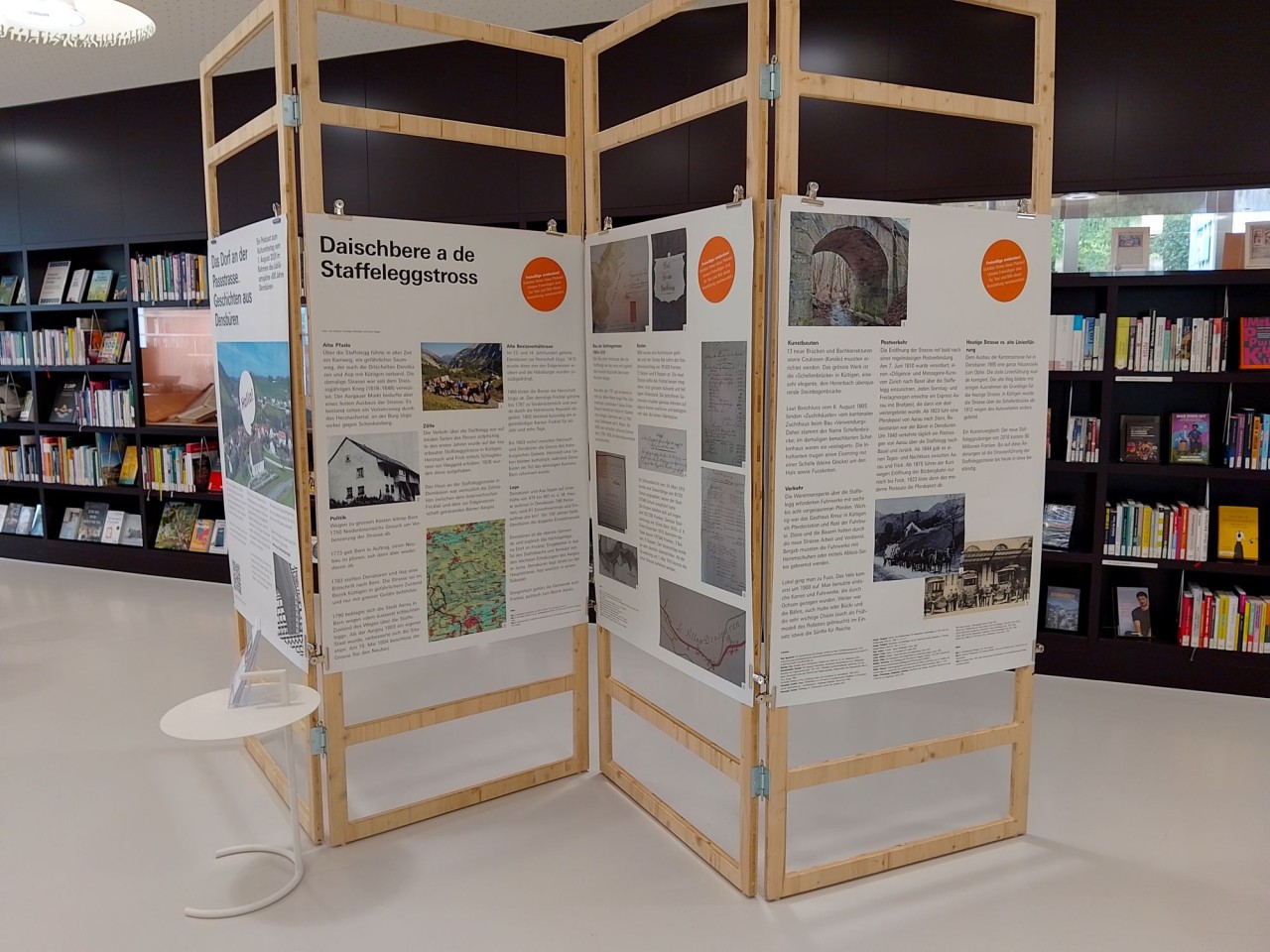 Eine Ausstellungstafel in einem offenen Holzrahmen mit Text- und Bildinformationen zur Geschichte der Staffeleggstrasse, vor einem Bücherregal in einer Bibliothek.