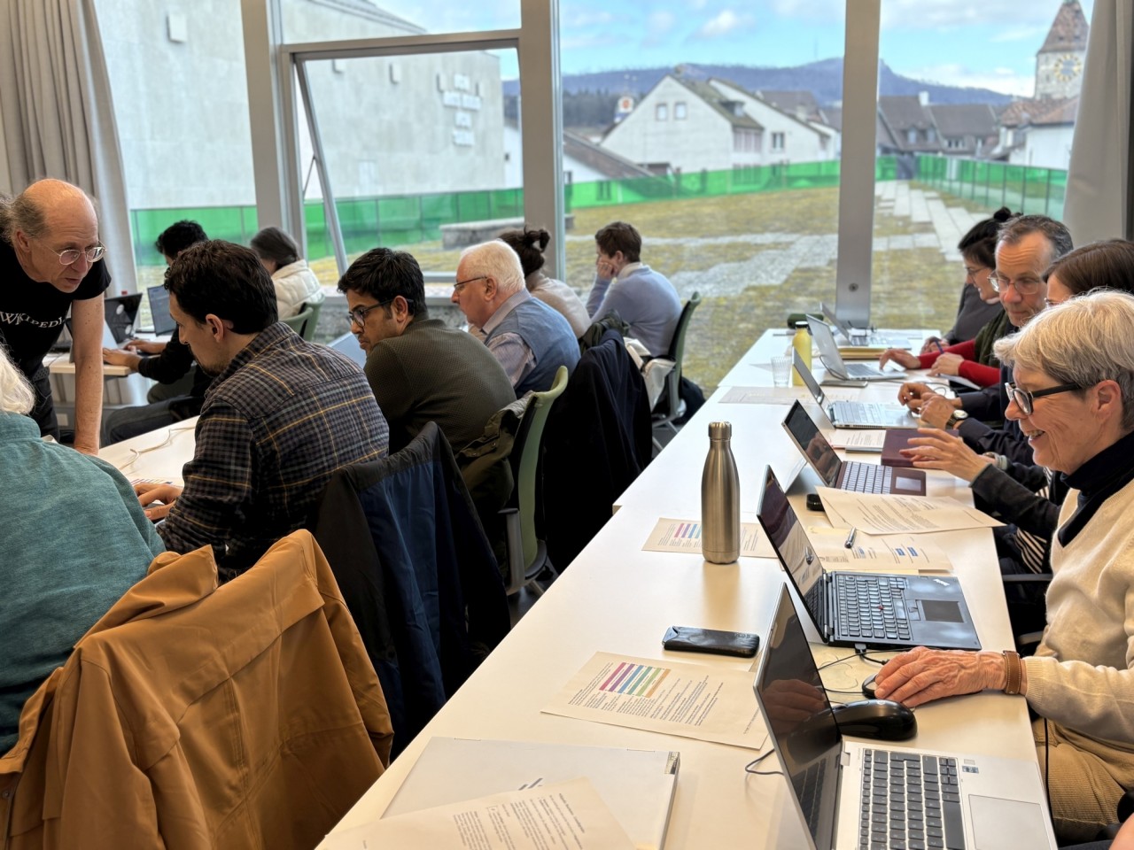 Menschen sitzen an einem langen Tisch mit Laptops und Dokumenten, vertieft in Diskussionen. Durch grosse Fenster ist eine städtische Umgebung mit Häusern und einem Kirchturm sichtbar.