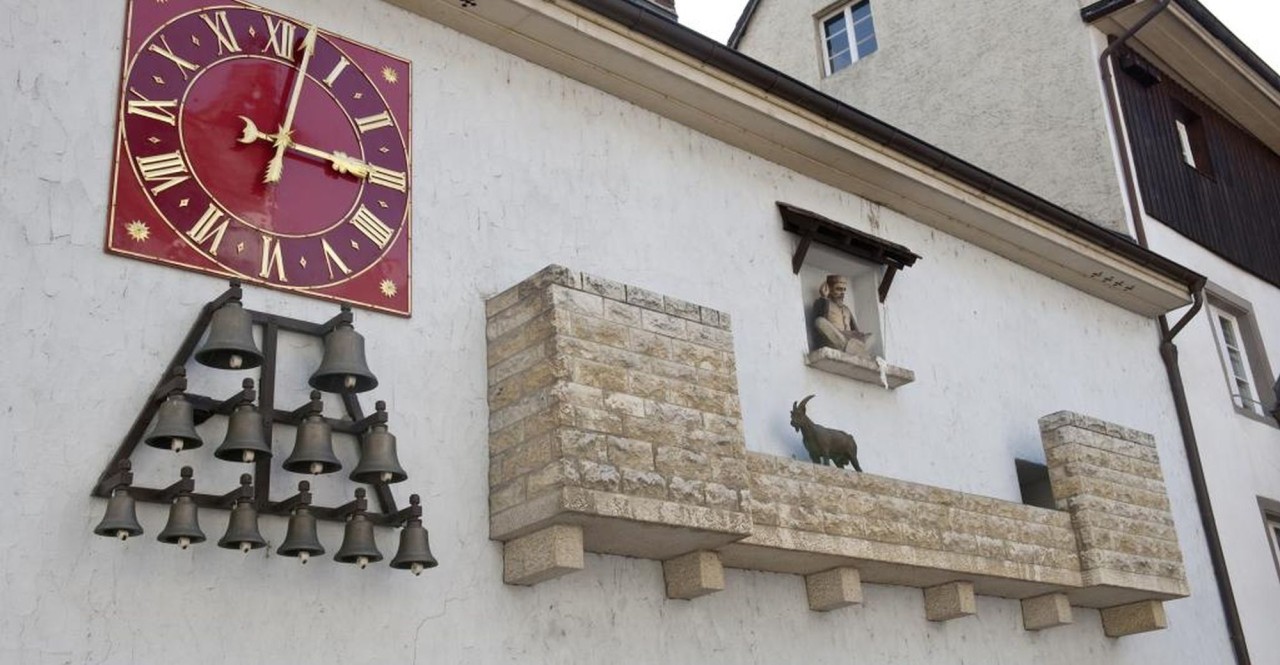 Rote Uhr mit römischen Zahlen neben einer Glockeninstallation an einer Wand, darunter eine Steinplattform mit Steinbockfigur und Statue in einer Aussparung.